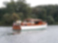 A restored wooden cruiser moving through calm waters, viewed from the stern.