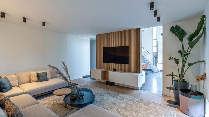 A modern living room with a beige sectional sofa, a round coffee table, and a large wall-mounted television. There are plants in the corners and natural light coming through a nearby window.