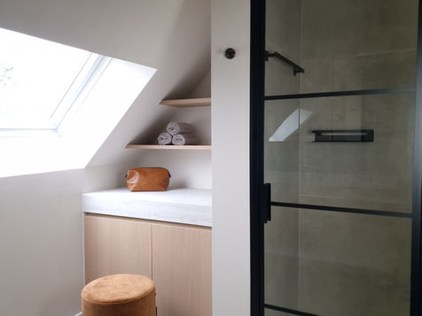 A modern bathroom with a sloped ceiling, featuring a glass shower enclosure, a wooden storage cabinet, and a small stool.