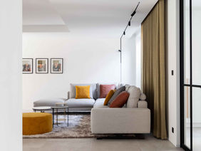 A modern living room with a light-colored sofa adorned with colorful cushions, a coffee table, and artwork on the walls. There is a cozy atmosphere created by natural light flowing through large windows and soft textures in the decor.
