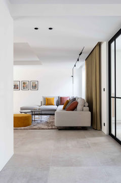 A modern living room with a light-colored sofa adorned with colorful cushions, a coffee table, and artwork on the walls. There is a cozy atmosphere created by natural light flowing through large windows and soft textures in the decor.