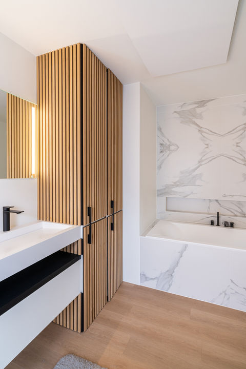 Een moderne badkamer met een houten kast met verticale latten, een wit aanrechtblad, een rechthoekige wastafel en een badkuip omgeven door marmeren wanden.