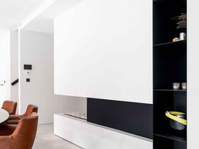 A modern interior space featuring a minimalistic design with a white wall unit, a black shelving unit, and a round dining table surrounded by brown chairs.