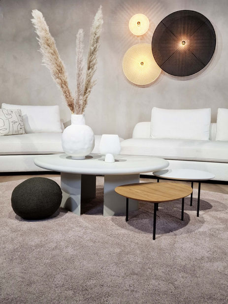 A modern living room featuring a concrete coffee table with a circular wooden side table beside it, a large white vase with dried pampas grass, and two round light fixtures on the wall. The room has a soft, neutral color palette with a plush area rug and white sofas.