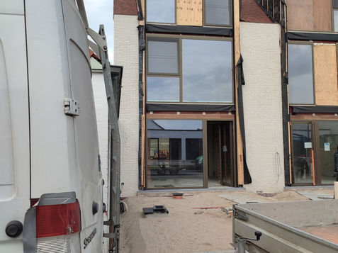 A construction site featuring a building under renovation, with wooden panels covering the windows and door. In the foreground, part of a van is visible, with a trailer attached, on a paved surface.