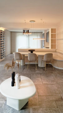 A modern dining area featuring a round coffee table with decorative items, four beige dining chairs around a long table, and a light-colored kitchen space with shelving and large windows.