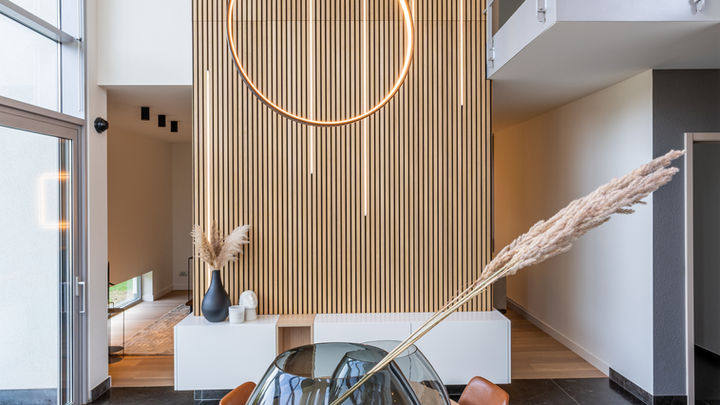 A modern dining area featuring a long wooden table with chairs, a large decorative vase in the center, and contemporary light fixtures hanging above. The walls are elegantly designed with a combination of natural and neutral tones.