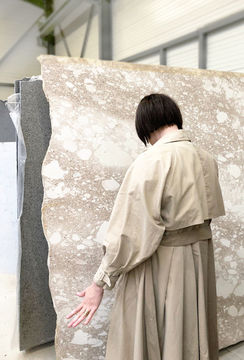 A person in a beige trench coat stands with their back to the camera, reaching out toward a large piece of textured stone or material. The setting appears to be an indoor studio or workspace.