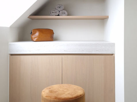 A modern bathroom space featuring a light wood cabinetry, a smooth white countertop, open shelves with decorative items, rolled towels, and a small round stool in an orange-brown tone.