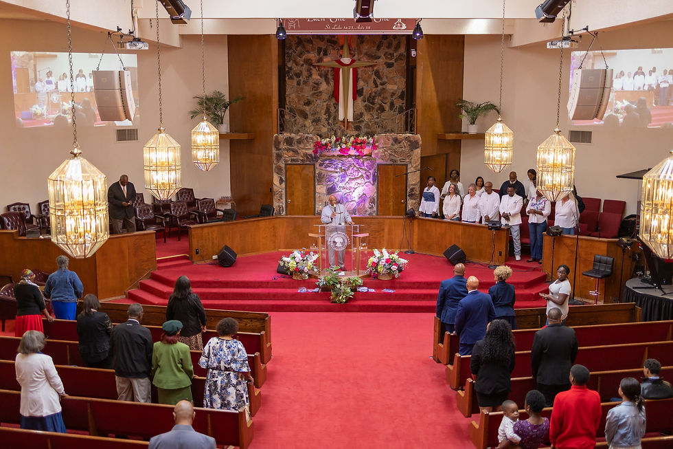 A pastor speaks at a church altar with a choir standing to the side. Congregation members stand in pews. The setting features red carpet and warm lighting.