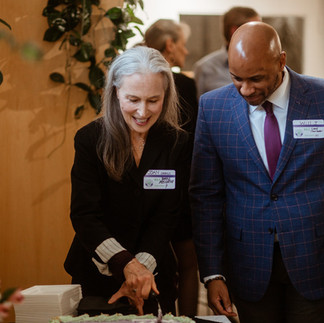 Two people smiling while cutting a large purple cake. They're indoors, wearing name tags. Green plants are in the background.