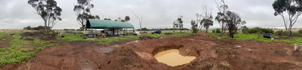 A caravan hides under a green shelter from the rain. A large hole has filled with mud in the foregound.