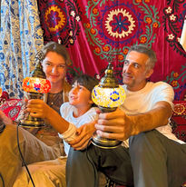 A family of three holding completed mosaic lamps, seated against a colorful Turkish textile background.