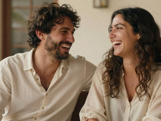 זוג יושב יחד בסלון, מחייך וצוחק זה עם זו, משדר קרבה רגשית, חיבור וביטחון בזוגיות.