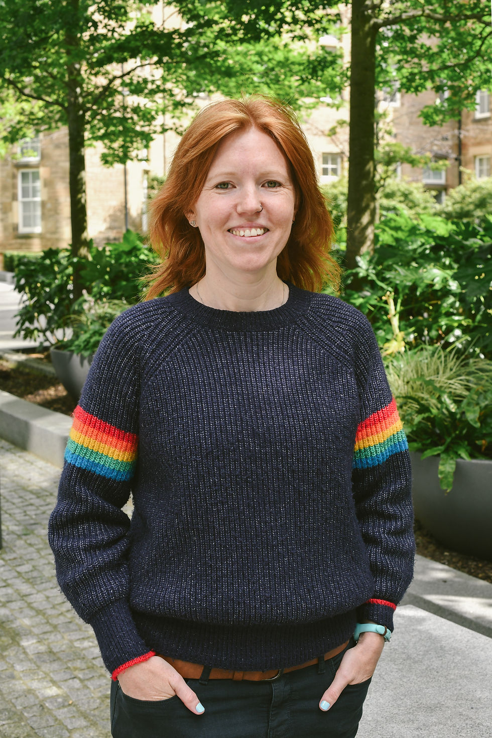 Photo of Chloe Foster smiling wearing a navy jumper with rainbow bands on the sleeves. Chloe is white and has ginger hair. Chloe is standing in front of green trees.