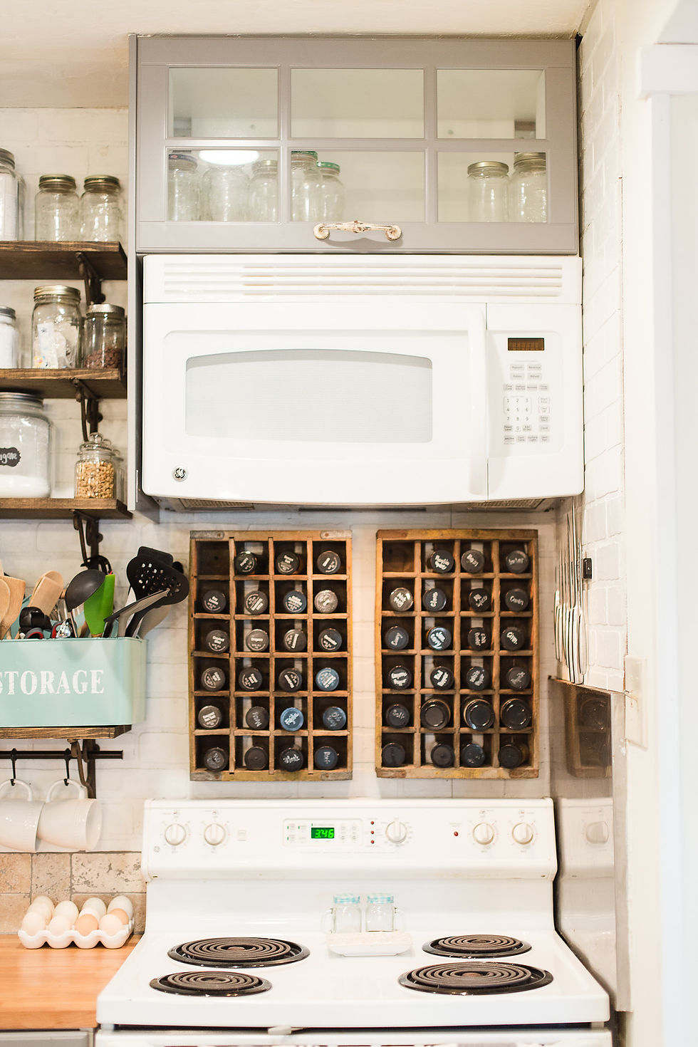 Kitchen scene with white microwave, spice rack, and utensils in a blue box labeled "STORAGE." Shelves hold jars. Cozy, organized vibe.