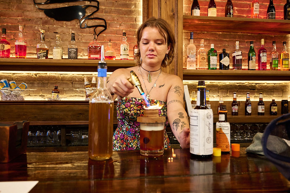 Bartender with tattoos uses a torch to ignite a cocktail on a wooden bar. Colorful shelves of bottles and brick walls create a cozy atmosphere.
