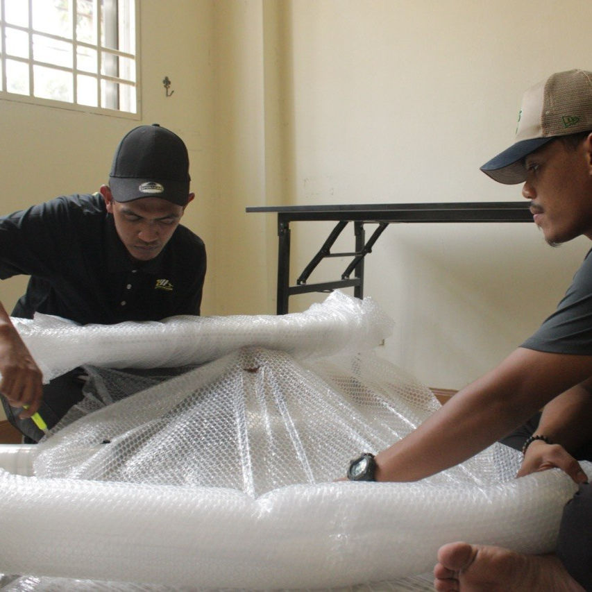 Two men in caps wrap a large object with bubble wrap in a bright room. A table is in the background, and the mood is focused.