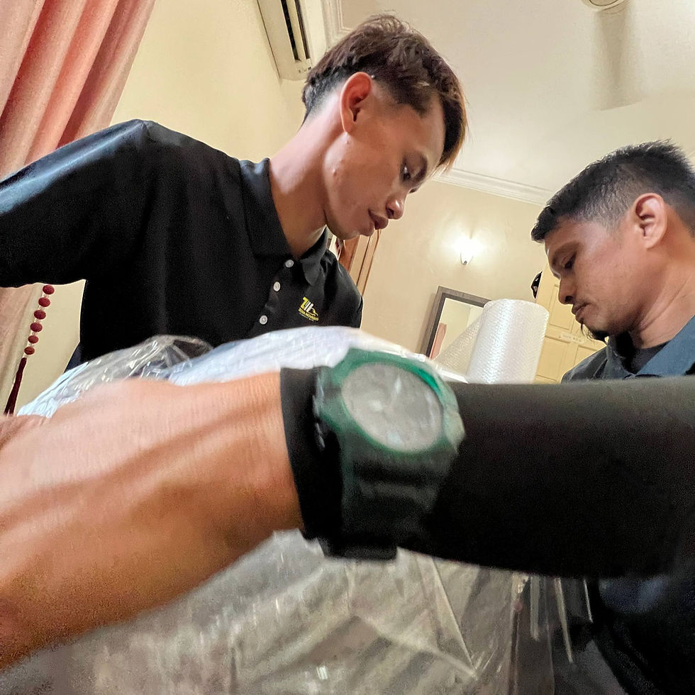 Two men lean over a wrapped package indoors, inspecting it closely; a wristwatch fills the foreground in a warm-lit room.
