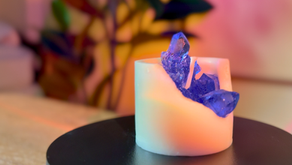 Homem segurando uma vela inspirada em pedra ametista, com acabamento artesanal que imita um geodo, sentado em uma mesa de madeira com decoração moderna ao fundo. A vela é feita com cera de coco e está pronta para ser encaixada em um copo de vidro.