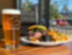 Beer glass with "Falling Sky" logo beside a burger with greens and fries on a plate. Bright, sunny day visible through window.