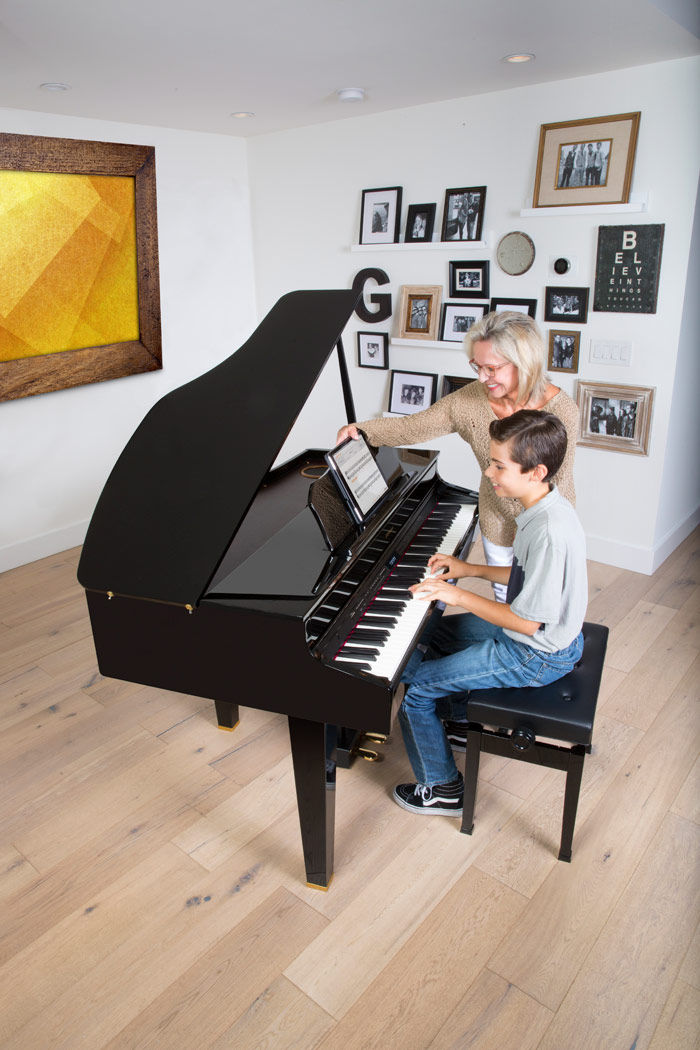 A boy plays piano with a woman guiding him in a room. The wall displays framed photos and art with a yellow painting. Mood is joyful.