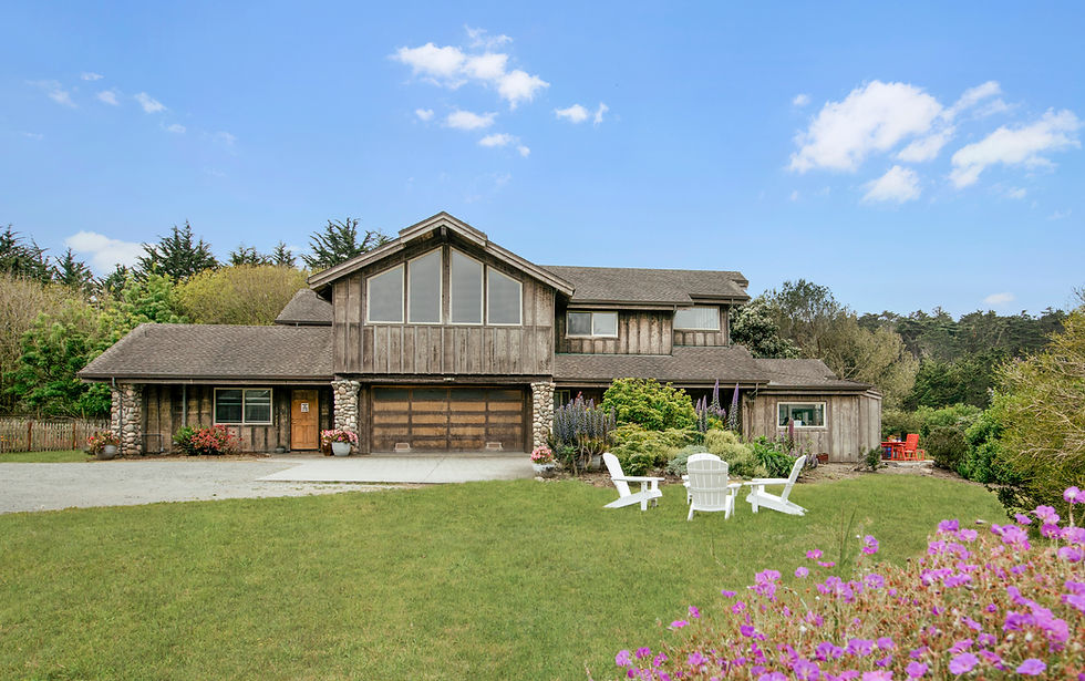 Large wooden home with a grassy lawn, white chairs, and trees.
