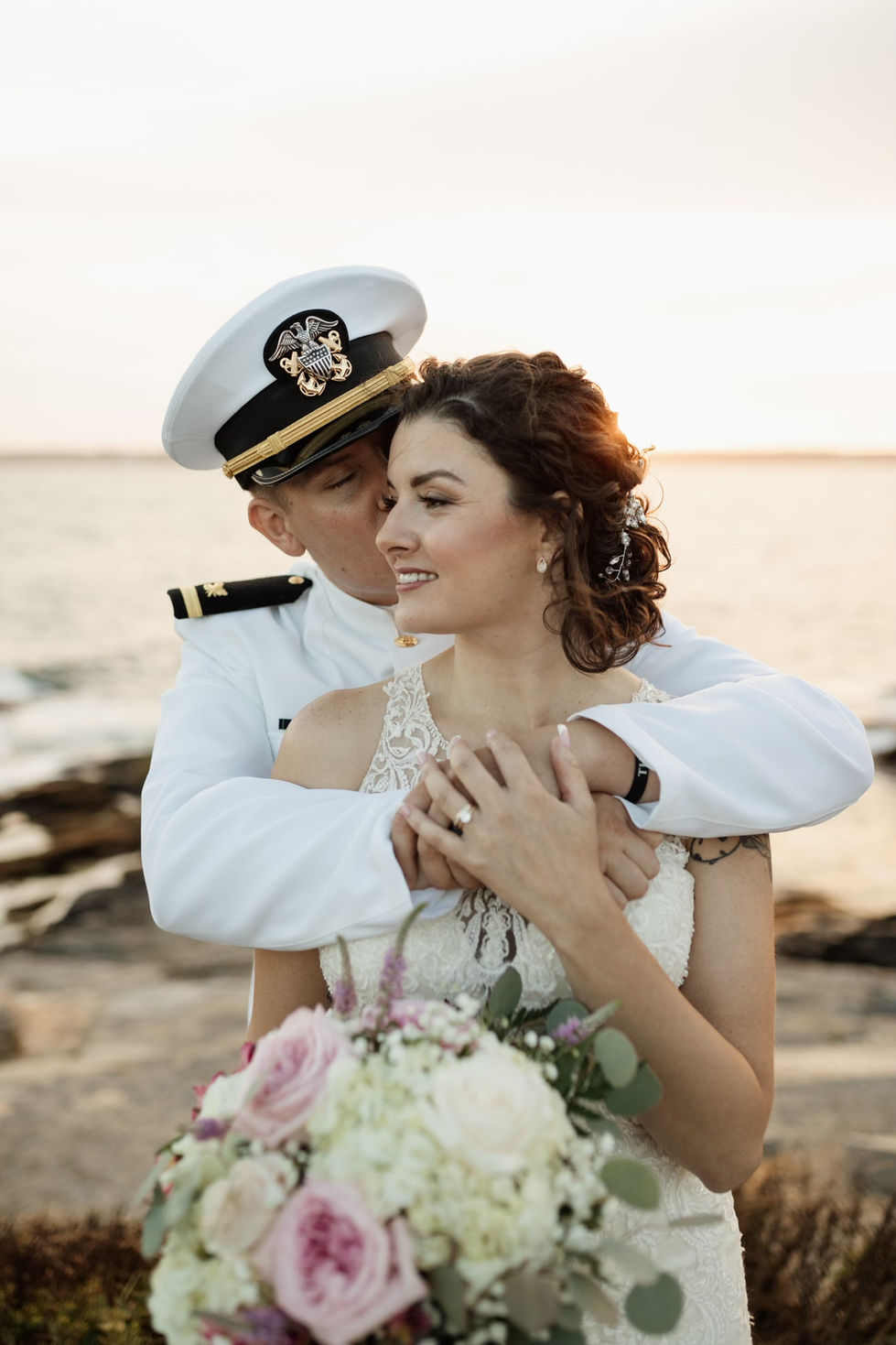 Bridal couple: naval groom embracing bride with bouquet at sunset