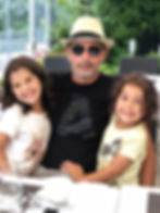 A man wearing a hat and sunglasses sitting at a restaurant table with two smiling young girls sitting on his lap.