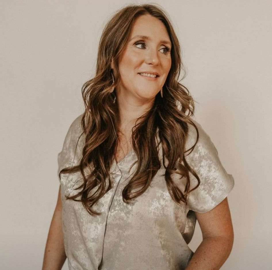 Woman with long wavy hair wearing a silver satin blouse, looking off to the side and smiling.