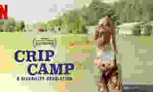 Crip Camp. A Disability Revolution. Shows a phto from the 70s. A young man with no shirt and a guitar over his shoulder pushes a teenage boy in a wheelchair. They are both smiling with a background of cabins, grass and trees. In the corner is the Netflix logo, and a logo indicating it was the Winner of the Audience award at the Sundance Film festival.