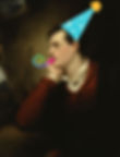 A colour portrait of a caucasian man with brown hair leaning his chin on his hand and facing to the side. There is a 2d sticker of a party hat on his head, and a multicoloured spiral to look like a party blower by his mouth