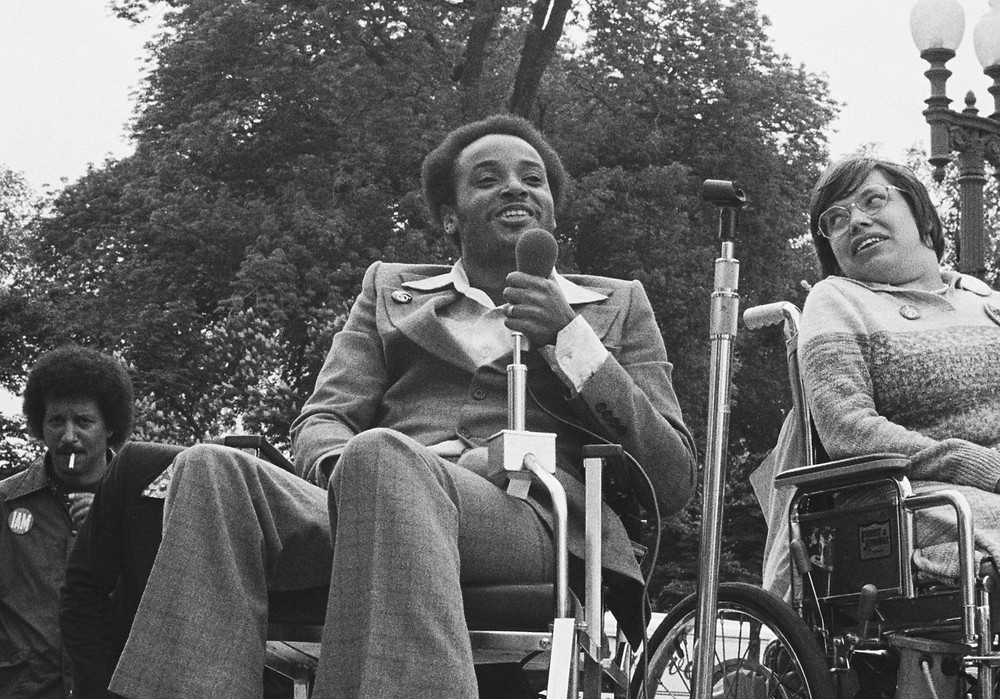 Black and white photo. A black man in a suit sits in his wheelchair holding a microphone to his mouth. On his right is a woman with glasses, also in a wheelchair facing slightly away from him and looking over her right shoulder at him