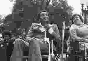 Black and white photo. A black man in a suit sits in his wheelchair holding a microphone to his mouth. On his right is a woman with glasses, also in a wheelchair facing slightly away from him and looking over her right shoulder at him