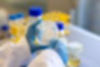 Lab worker in blue gloves holds a petri dish with white streaks. Blurred yellow test tube rack and bottles in the background.