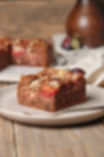 Zwetschgen-Käsekuchenstück auf Teller, im Hintergrund unscharf ein zweites Stück und eine weiße Vase mit Trockenblumen.