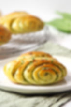 Herzhaftes Franzbrötchen aus Hefeteig mit Bärlauch auf weißem Teller, im Hintergrund unscharf weitere goldbraune Brötchen.