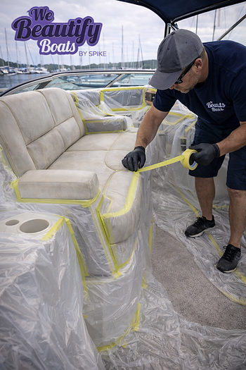 Technician from We Beautify Boats by Spike carefully applying precision masking and plastic sheeting to protect surrounding surfaces before vinyl recoloring inside a boat cockpit at a marina.