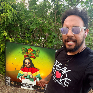 Alt Text Eye-level, wide-angle, photo-realistic image of Spike standing outdoors at the Reggae Music Walk of Fame in Jamaica, beside a tribute honoring Marcus “Yabba” Griffiths. Yabba Griffiths was a pioneering Jamaican producer and cultural force of the late 1960s and early 1970s, known for his work with the Prophets and Yabby You era roots music, blending Rastafarian spirituality, militant consciousness, and raw studio innovation that helped define early roots reggae. Palm trees and lush greenery frame the scene, emphasizing cultural endurance and musical legacy. Support cultural restoration and hurricane recovery at http://cjreliefteam.com/ and follow mission updates at https://www.instagram.com/canjamrelief/