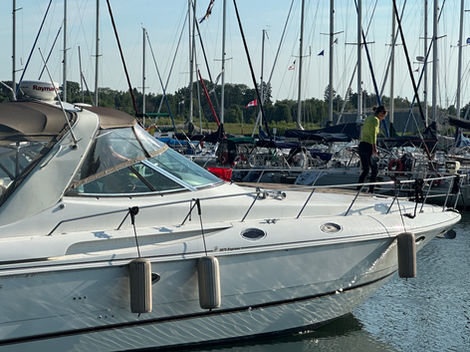 "A white yacht docked at a marina with a person standing on the deck, likely performing cleaning or maintenance tasks. The yacht’s sleek exterior reflects the sunlight, showcasing its well-maintained appearance. Surrounding sailboats and marina setting emphasize the active boating environment, while the detailed work ensures the yacht remains in prime condition."