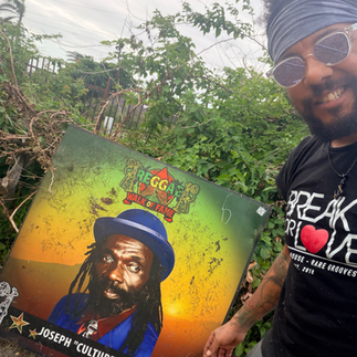 Alt Text Eye-level, wide-angle, photo-realistic image of Spike standing outdoors at the Reggae Music Walk of Fame in Jamaica, beside a tribute honoring Joseph “Culture” Hill. Joseph Hill was the lead singer and songwriter of the legendary roots reggae group Culture, whose music became synonymous with Rastafarian consciousness and spiritual resistance. Iconic songs such as “Two Sevens Clash,” “International Herb,” and “Natty Never Get Weary” remain foundational works in reggae history, shaping the sound and message of roots reggae worldwide. Palm trees and lush tropical greenery frame the scene, symbolizing prophecy, endurance, and lasting cultural impact. Support cultural restoration and hurricane recovery at http://cjreliefteam.com/ and follow updates at https://www.instagram.com/canjamrelief/