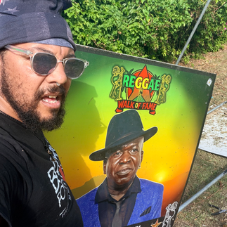 Eye-level, wide-angle, photo-realistic image of Spike standing outdoors at the Reggae Music Walk of Fame in Jamaica, beside a tribute honoring Barrington Levy. Barrington Levy is a legendary Jamaican reggae and dancehall vocalist known for his distinctive high tenor voice and crossover appeal between roots reggae and early dancehall. His iconic songs, including “Here I Come,” “Under Mi Sensi,” and “Murderer,” remain global anthems and have been widely sampled across hip-hop and modern music. Palm trees and lush tropical greenery frame the scene, symbolizing longevity, influence, and reggae’s evolving sound. Support cultural restoration and hurricane recovery at http://cjreliefteam.com/ and follow updates at https://www.instagram.com/canjamrelief/