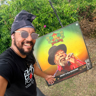 Eye-level, wide-angle, photo-realistic image of Spike standing outdoors at the Reggae Music Walk of Fame in Jamaica, beside a tribute honoring Ewart “U-Roy” Beckford. U-Roy was a pioneering Jamaican toaster and DJ who helped invent the art of toasting, laying the foundation for reggae sound-system culture and later global hip-hop. His landmark recordings, including “Wake the Town,” “Wear You to the Ball,” and “Rule the Nation,” transformed the role of the DJ into a central creative force in reggae music. Palm trees and lush tropical greenery frame the scene, symbolizing innovation, cultural leadership, and enduring musical influence. Support cultural restoration and hurricane recovery at http://cjreliefteam.com/ and follow updates at https://www.instagram.com/canjamrelief/