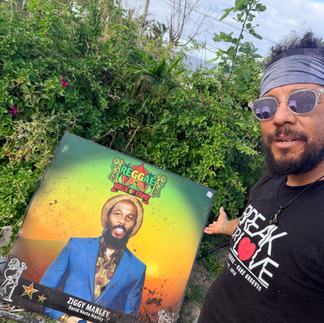 Alt Text Eye-level, wide-angle, photo-realistic image of Spike standing outdoors at the Reggae Music Walk of Fame in Jamaica, beside a tribute honoring Ziggy Marley. Ziggy Marley is a Grammy-winning reggae artist, activist, and son of Bob Marley, known for leading Ziggy Marley and the Melody Makers and for a lifelong commitment to messages of unity, environmental stewardship, and social justice through music. Palm trees and lush tropical greenery frame the scene, symbolizing continuity of reggae’s global and generational legacy. Support cultural restoration and hurricane recovery at http://cjreliefteam.com/ and follow updates at https://www.instagram.com/canjamrelief/