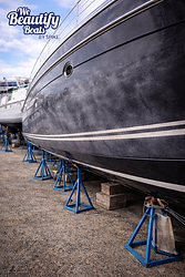 Photo-realistic image of a large dark-colored boat hull elevated on blue jack stands in a boatyard, showing severe oxidation and chalking across the topsides. The gelcoat appears dull, hazy, and uneven with visible patchy discoloration, indicating heavy surface degradation. White accent stripes run along the hull, contrasting with the faded dark finish. The surrounding gravel yard and nearby vessels reinforce the working marina environment. The We Beautify Boats by Spike logo is positioned in the upper left corner. The image represents a pre-restoration condition requiring Level 4 hull polishing to remove severe oxidation, restore depth of color, and achieve a smooth, high-gloss finish. Service by We Beautify Boats by Spike, Boat Detailing, Toronto, Ontario, Canada.