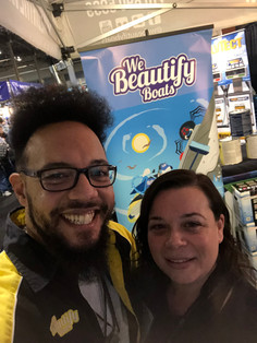 Shane "Spike" Desloges, founder of We Beautify Boats, smiles widely alongside a colleague at a boating event. Spike wears a black and yellow jacket with the We Beautify Boats logo, while his colleague smiles warmly beside him. Behind them, a vibrant We Beautify Boats banner featuring a stylized wave design and the company’s branding creates a professional and inviting atmosphere. Image credit: webeautifyboats.com.