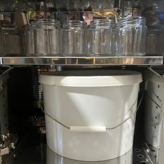 A 30lb honey bucket and some glass jars being warmed in a honey warming cabinet