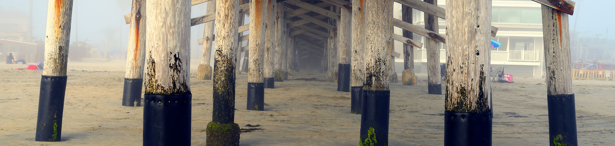 View beneath a pier with wooden beams overhead, darker support posts, and sand and ocean water in the background.
