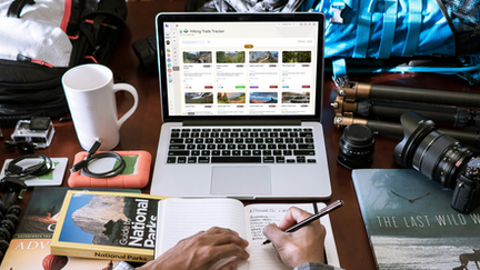 Person planning hiking adventures using Hylark’s Hiking Trails Tracker on a laptop, surrounded by travel gear, camera, and notebooks — organizing hiking routes and outdoor hobbies in one digital workspace.