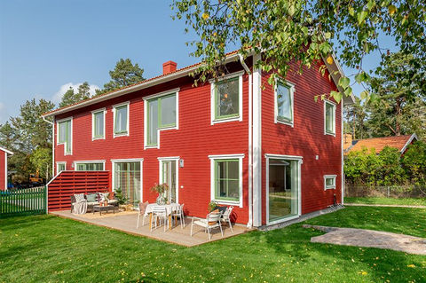 Traditional two-story house with a red wooden facade and white trim around the windows and doors. The house features large windows and an outdoor patio area with seating. A green lawn surrounds the property, with trees and neighboring houses in the background under a clear sky.